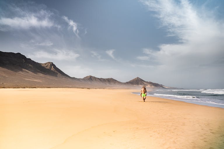 A person walking on a pristine beach in Fuerteventura with rugged mountains in the background.